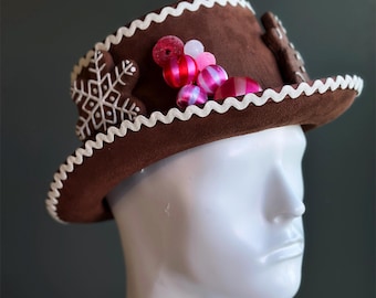 Peut inclure: Un chapeau haut de forme marron avec une bordure festonnée blanche et des décorations. Le chapeau présente un motif de flocon de neige et de bonhomme en pain d'épices, ainsi que des boules roses et rouges. Le chapeau est présenté sur une tête de mannequin blanche.