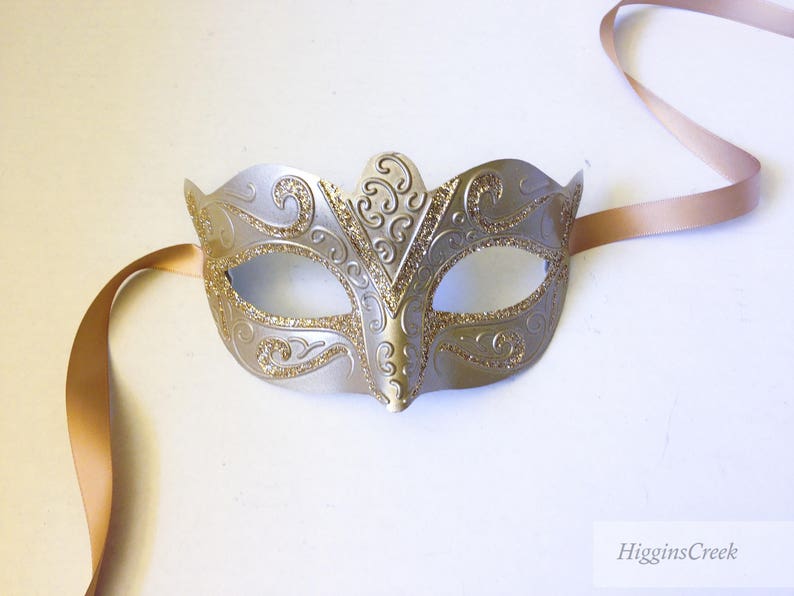 May include: A silver masquerade mask with gold glitter accents and a delicate, ornate design. The mask is attached to a long, thin, gold ribbon.
