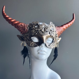 Puede incluir: Una máscara de mascarada decorativa con detalles florales en bronce y oro, cuernos curvos y plumas negras colgantes. La máscara presenta el texto "MASQUERADE STORE .COM" en la parte superior. Diseñada para un baile de máscaras o fiesta de disfraces.
