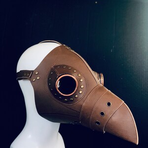 Pode incluir: Uma m&aacute;scara de m&eacute;dico da peste de couro marrom com um grande olho redondo e detalhes em metal. A m&aacute;scara est&aacute; presa a uma cabe&ccedil;a de manequim branca.