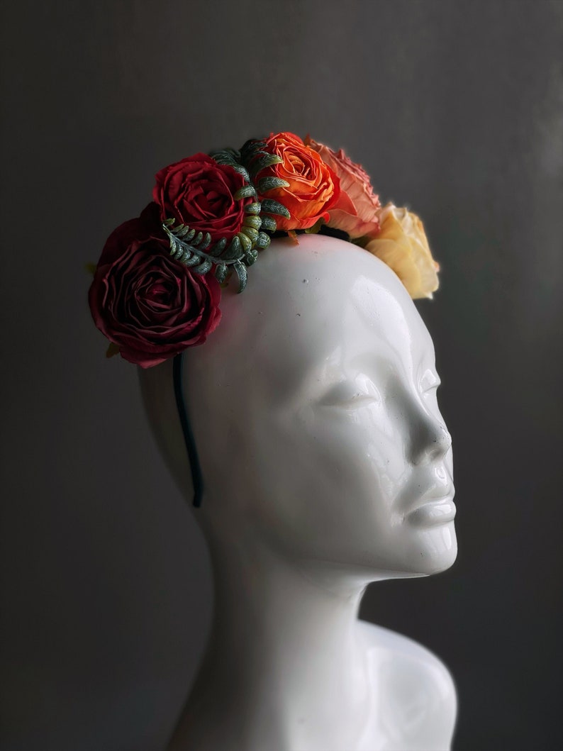 May include: A floral headband featuring red, orange and cream silk flowers. The headband is made from dark green wire and is decorated with green leaves.