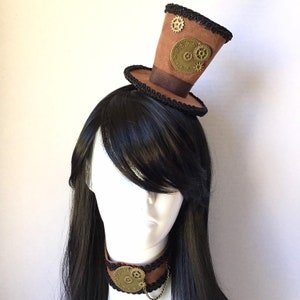 Op de afbeelding: Een bruine hoge hoed met een zwarte rand en gouden tandwielen en wijzerplaataccenten. De hoed wordt gedragen door een mannequin met lang zwart haar.