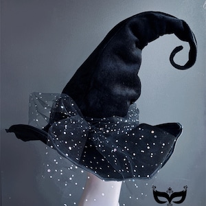 Puede incluir: Un sombrero de bruja negro con acabado de terciopelo y punta rizada. El sombrero presenta un ala ancha con una capa de tela transparente con lunares. Un lazo adorna la base. El logotipo de Masquerade Store está en la esquina inferior derecha.