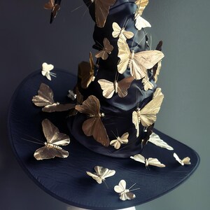 May include: A black witch hat adorned with numerous gold-colored butterfly embellishments. The hat features a wide brim and a tall, pointed crown, creating a whimsical and elegant accessory. The Masquerade Store logo is visible.