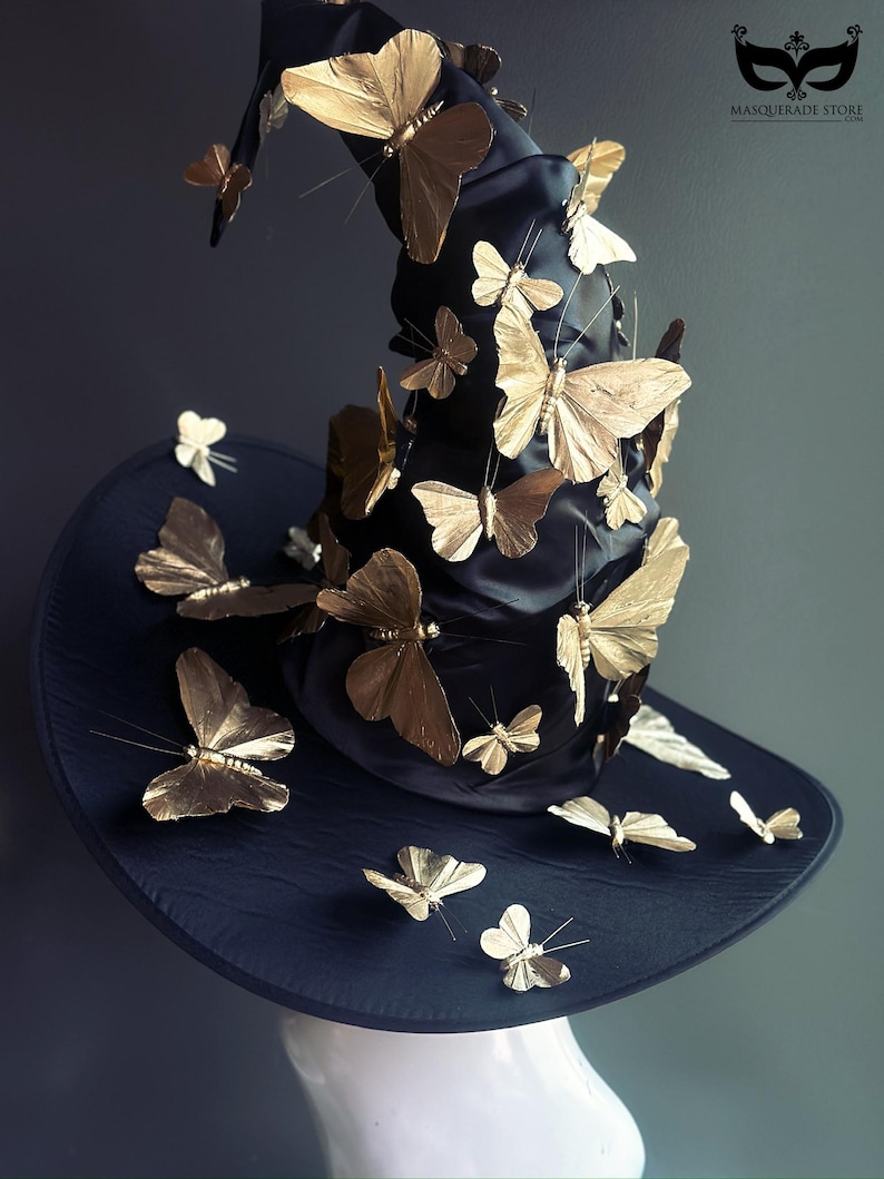 May include: A black witch hat adorned with numerous gold-colored butterfly embellishments. The hat features a wide brim and a tall, pointed crown, creating a whimsical and elegant accessory. The Masquerade Store logo is visible.