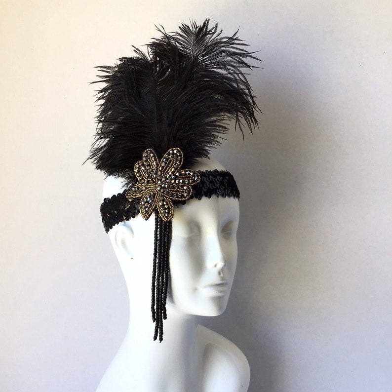 May include: A black headband with a black feather and a gold and black beaded flower. The headband is adorned with black sequins.