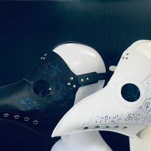 Puede incluir: Dos m&aacute;scaras de m&eacute;dico de la peste, una negra y otra blanca, con dise&ntilde;os florales azules y morados y agujeros para los ojos. Las m&aacute;scaras est&aacute;n hechas de cuero y tienen un pico largo.