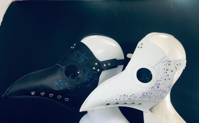 Puede incluir: Dos m&aacute;scaras de m&eacute;dico de la peste, una negra y otra blanca, con dise&ntilde;os florales azules y morados y agujeros para los ojos. Las m&aacute;scaras est&aacute;n hechas de cuero y tienen un pico largo.