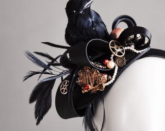 May include: A black fascinator headband featuring a prominent black feathered bird, black satin ribbon loops, metallic gear-shaped charms, small white faux pearls, and an ornate gold-tone brooch with a central red stone accent.
