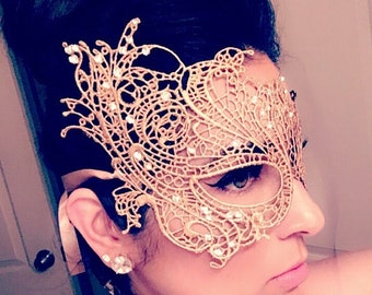 May include: A gold lace masquerade mask with rhinestones. The mask covers the eyes and part of the nose. The mask is worn by a person with dark hair.