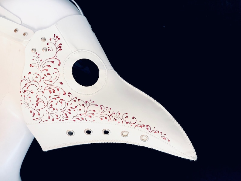 Puede incluir: M&aacute;scara blanca de m&eacute;dico de la peste con un pico largo y un dise&ntilde;o floral rojo. La m&aacute;scara tiene un agujero redondo para el ojo y una serie de ojales a lo largo del pico.
