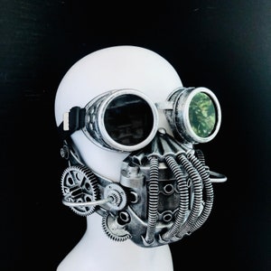 May include: Steampunk mask with silver goggles and a detailed metallic face covering. The mask features gears, tubes, and a textured finish, creating a futuristic, industrial aesthetic. The goggles have dark lenses and a black strap.