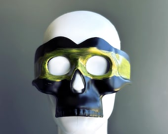 May include: A black and gold skull mask with eye holes and a nose opening. The mask is worn on a white mannequin head. The mask has a gold band across the forehead and is from the Masquerade Store.