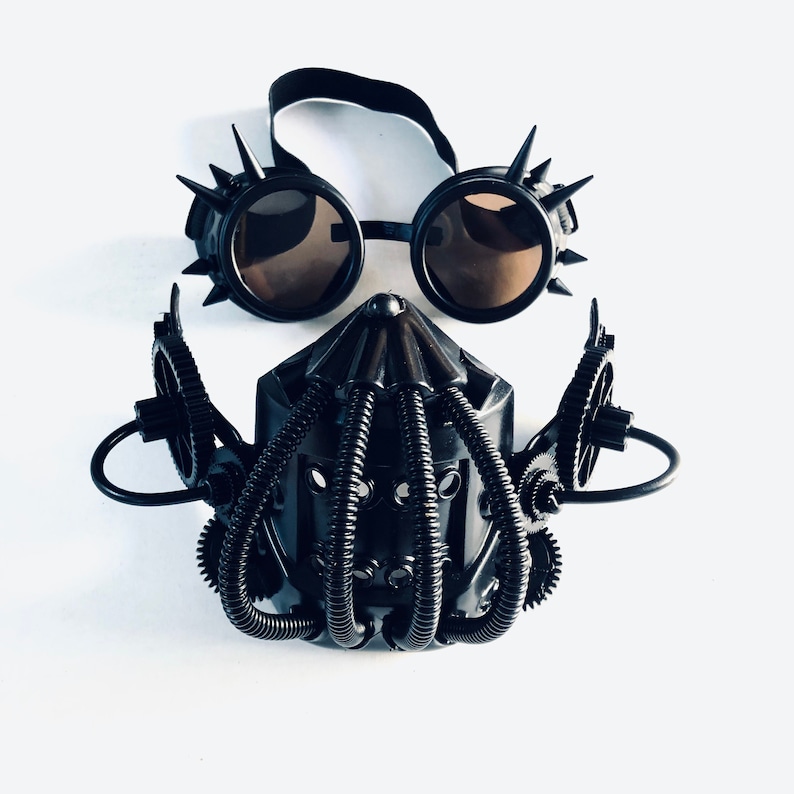 May include: A black steampunk mask and goggles set against a white backdrop. The goggles have spiked rims and tinted lenses. The mask features a cage-like design with gear and tube details, creating a post-apocalyptic aesthetic.