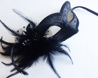 May include: A black masquerade mask with intricate details and a black feather trim. The mask has a delicate lace design and a silver glitter finish.