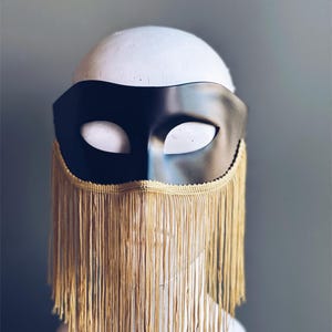 May include: A black masquerade mask with gold fringe. The mask has a classic design with eye holes and a curved shape.