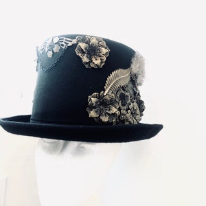 May include: A black top hat with a wide brim and a tall crown, decorated with gold-colored metal flowers and leaves. The hat is on a white mannequin head.