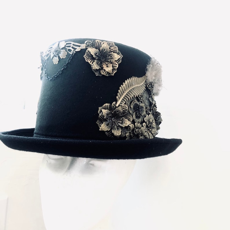 May include: A black top hat with a wide brim and a tall crown, decorated with gold-colored metal flowers and leaves. The hat is on a white mannequin head.