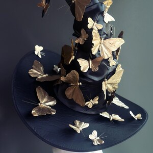 May include: A black witch hat adorned with numerous gold-colored butterfly decorations. The hat has a wide brim and a tall, pointed crown. The butterflies are of varying sizes and are attached to the hat.