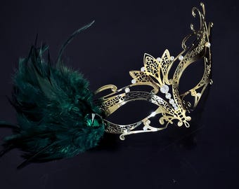 May include: A gold masquerade mask with intricate cutouts and rhinestones. The mask has a green feather attached to the side.