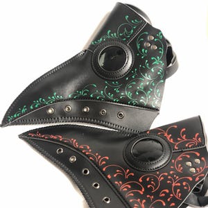 May include: Two black leather plague doctor masks with a decorative floral pattern. One mask has a green pattern and the other has a red pattern.