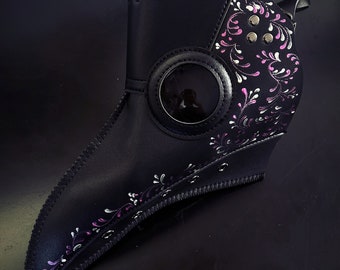 Op de afbeelding: Een zwart leren pestdoktersmasker met een lange snavel en een rond oogstuk. Het masker is versierd met een roze en wit bloemenontwerp.