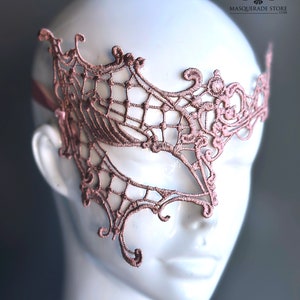 May include: A delicate, blush-colored lace masquerade mask with intricate floral and web-like patterns. The mask covers one eye and is designed to be worn over the face.