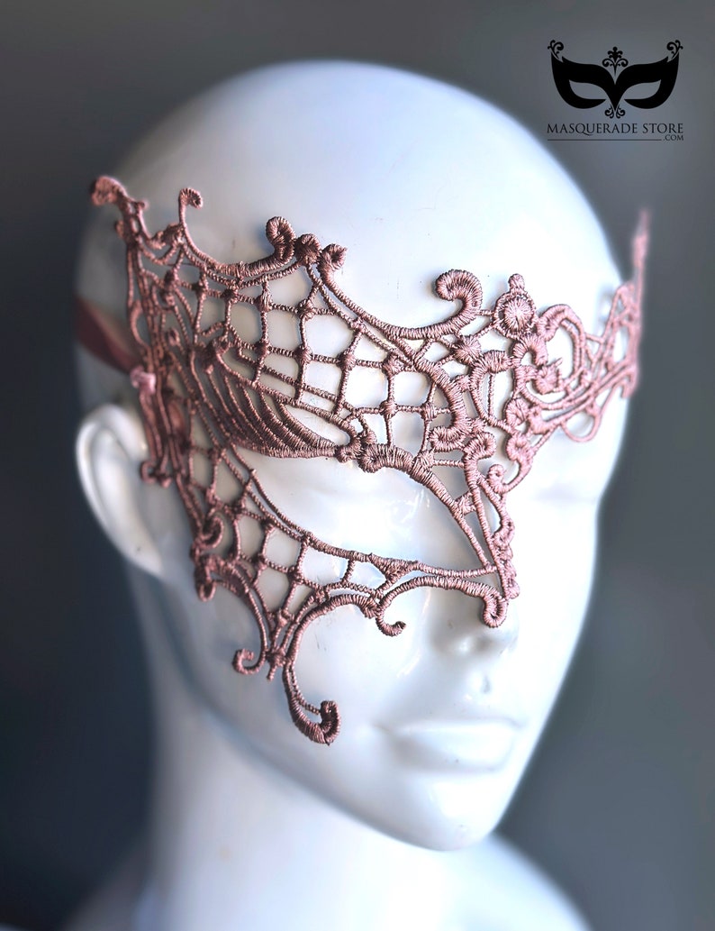 May include: A delicate, blush-colored lace masquerade mask with intricate floral and web-like patterns. The mask covers one eye and is designed to be worn over the face.