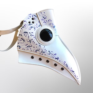 May include: White leather plague doctor mask with a purple and blue floral design. The mask has a large round eye hole and a long beak.