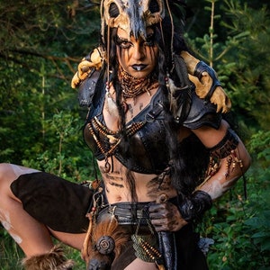 May include: A woman wearing a costume with a skull headdress, leather armor, and a fur skirt. She is holding a knife in her right hand and has white paint on her face and arms.