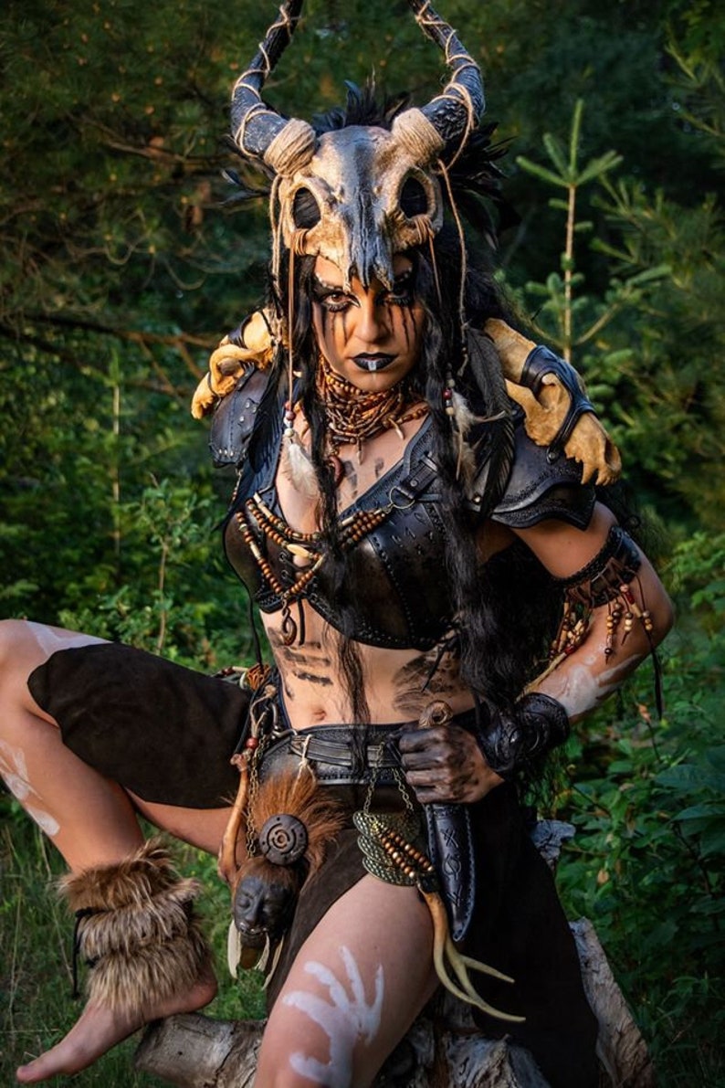May include: A woman wearing a costume with a skull headdress, leather armor, and a fur skirt. She is holding a knife in her right hand and has white paint on her face and arms.