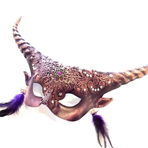 May include: A brown and purple masquerade mask with a horn-like design. The mask features intricate lace detailing, sparkling rhinestones, and a central purple jewel. Purple feathers dangle from the bottom, adding a touch of elegance.