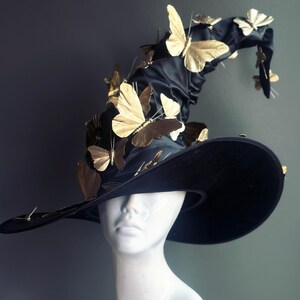May include: A black witch hat with a wide brim, adorned with numerous gold butterfly embellishments. The hat is displayed on a white mannequin head, showcasing its dramatic shape and decorative details. The hat's design suggests a masquerade or costume theme.