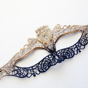May include: A black and gold lace masquerade mask with intricate details. The mask is tied with a black ribbon.