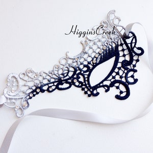 Peut inclure: Un masque de bal masqu&eacute; en dentelle noire et argent&eacute;e avec des strass et un ruban de satin blanc.