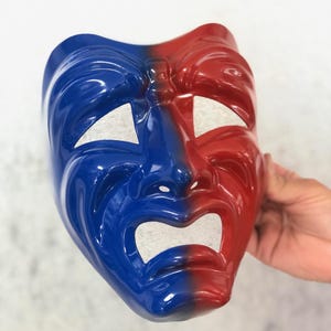 May include: A theatre mask divided into blue and red sections. The mask features eye cut-outs and a wide, open mouth. It represents drama and performance, a classic theatrical symbol.