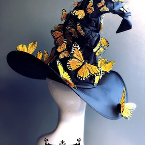 Pu&ograve; includere: Un cappello da strega nero con una corona alta e appuntita decorata con numerosi accenti di farfalle gialle e arancioni. Il cappello &egrave; esposto su un manichino bianco.