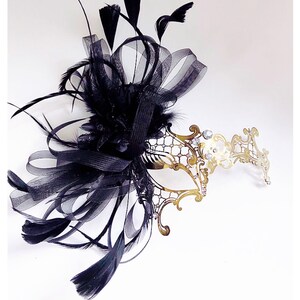 May include: A gold masquerade mask with a delicate, intricate design. The mask is adorned with rhinestones and has a black feather and ribbon embellishment.