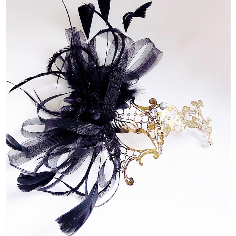 May include: A gold masquerade mask with a delicate, intricate design. The mask is adorned with rhinestones and has a black feather and ribbon embellishment.