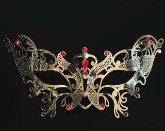 Può includere: Una maschera dorata con intricati disegni a filigrana e accenti di gioielli rossi. La maschera ha una forma simmetrica a farfalla ed è posta su uno sfondo nero.