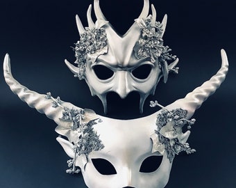 Puede incluir: Dos máscaras de plata para el baile de máscaras con intrincados detalles florales. Una máscara tiene un diseño demoníaco con cuernos, mientras que la otra tiene un diseño más delicado y femenino.