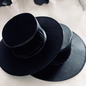 Pode incluir: Dois chap&eacute;us de feltro preto empilhados um em cima do outro. O chap&eacute;u de cima tem uma coroa alta e cil&iacute;ndrica e uma aba estreita. O chap&eacute;u de baixo tem uma aba mais larga.