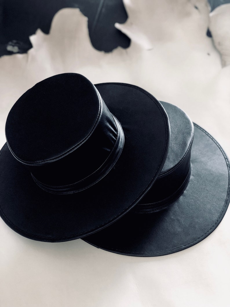 Pode incluir: Dois chap&eacute;us de feltro preto empilhados um em cima do outro. O chap&eacute;u de cima tem uma coroa alta e cil&iacute;ndrica e uma aba estreita. O chap&eacute;u de baixo tem uma aba mais larga.