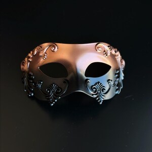 Op de afbeelding: Een metallic bronzen maskerademasker met ingewikkelde, donkere filigraan details. Het masker heeft twee ooggaten en is ontworpen met een gebogen vorm. De decoratieve elementen zijn symmetrisch, wat bijdraagt aan de elegante uitstraling van het masker.