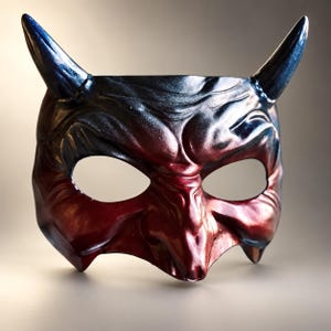 May include: A devil mask featuring two prominent horns, characterized by a metallic gradient finish that transitions from a deep, dark blue at the top to a vibrant, glossy red across the lower facial features, including the nose and cheekbones.