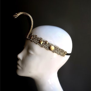 May include: A gold-colored headband with a snake design. The headband features an ornate, filigree pattern with decorative jewels. The snake rises above the head, creating a unique and eye-catching accessory. The headband is on a white mannequin head against a dark background.