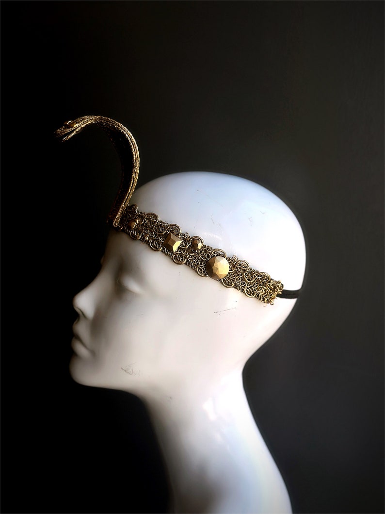 May include: A gold-colored headband with a snake design. The headband features an ornate, filigree pattern with decorative jewels. The snake rises above the head, creating a unique and eye-catching accessory. The headband is on a white mannequin head against a dark background.