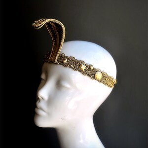 May include: A gold-colored headband with a cobra design. The cobra rises from the center of the headpiece. The headband features an ornate, filigree design with square and round embellishments. The headpiece is displayed on a white mannequin head against a dark background.