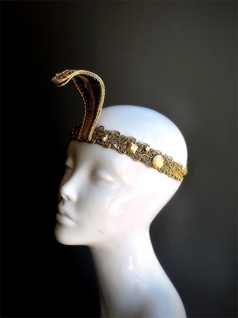 May include: A gold-colored headband with a cobra design. The cobra rises from the center of the headpiece. The headband features an ornate, filigree design with square and round embellishments. The headpiece is displayed on a white mannequin head against a dark background.