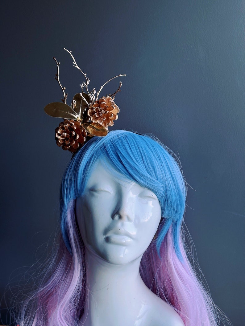 May include: A gold and brown pine cone hairpiece with twigs and leaves. The hairpiece is worn on a mannequin with blue and pink hair.